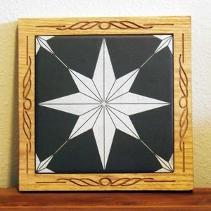 May include: Square wooden framed tile featuring a white starburst design on a black background. The frame is light brown with decorative carved accents. The tile is likely for home decor.