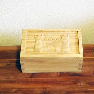 May include: A light-colored wooden box with a carved castle design on the lid. The box is rectangular and sits on a wooden surface. The castle design includes two towers and a central arched doorway.