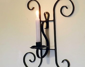 May include: Black wrought iron wall sconce with a lit white candle. The sconce features a decorative design with curved and scrolled details. The candle is centered, emitting a warm light. A classic home decor item.