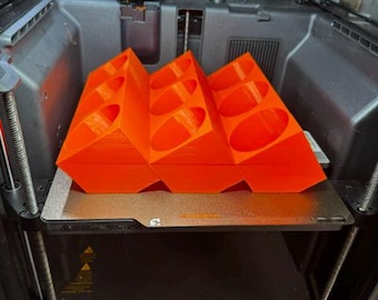 Könnte beinhalten: Ein leuchtend orangefarbener 3D-gedruckter Organizer mit neun schrägen zylindrischen Fächern in einem Drei-mal-drei-Raster. Das Objekt steht auf der metallischen Druckplatte innerhalb eines 3D-Druckers und weist ein geometrisches Design auf.