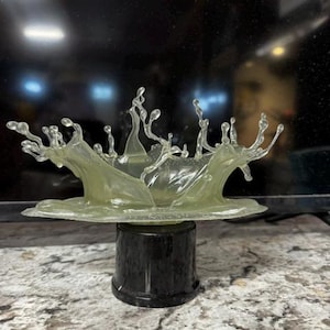 May include: A translucent, pale yellow sculpture depicting a frozen liquid splash with multiple upward-reaching droplets. The sculpture is mounted on a solid, cylindrical black base, creating a contrast between the clear top and dark bottom.