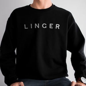 May include: Black crew neck sweatshirt with the word "LINGER" printed in silver across the chest. The sweatshirt is a solid black color and has a classic, minimalist design. The text is centered and appears in a sans-serif font.