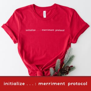 Może przedstawiać: Jasnoczerwona koszulka z białym napisem "initialize ....: merriment protocol". Koszulka jest zawiązana na dole i ma podwinięte rękawy. Gałązka iglasta i szyszka umieszczone są na dole.