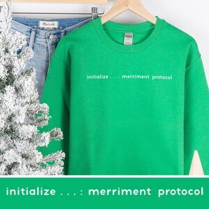 Może przedstawiać: Jasnozielona bluza z białym napisem "initialize ...: merriment protocol". Bluza jest prezentowana z niebieskimi dżinsami i białym drzewkiem świątecznym. Odzież wykonana jest z miękkiego materiału.