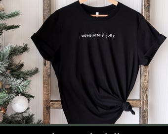 May include: Black t-shirt with the phrase "adequately jolly" in white, displayed on a wooden hanger. The shirt is knotted at the bottom. A Christmas tree and a wooden ladder are in the background.