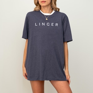 May include: A navy blue t-shirt with the word "LINGER" printed in white across the chest. The shirt has short sleeves and a white crew neck collar. The model is wearing a gold necklace.