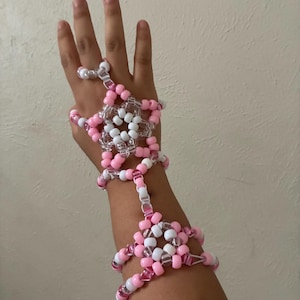 Könnte beinhalten: Ein handgefertigtes Perlen-Handgeschirr aus rosa, weißen und transparenten Perlen. Das Geschirr hat ein Blumenmuster auf dem Handrücken und verbindet sich mit einem Armband. Die Perlen sind mit durchsichtigem Faden aufgefädelt.