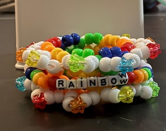 Könnte beinhalten: Ein Stapel bunter Perlenarmbänder. Die Armbänder sind weiß, rot, orange, gelb, grün und blau. Ein Armband schreibt das Wort "RAINBOW" mit weißen Buchstabenperlen. Einige Perlen sind sternförmig.