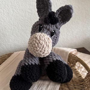 Könnte beinhalten: Ein handgehäkeltes Esel-Plüschtier in Grau-, Schwarz- und Beigetönen. Der Esel hat große Ohren, eine flauschige Schnauze und schwarze Hufe. Das Stofftier sitzt auf einem hellen Stoff.