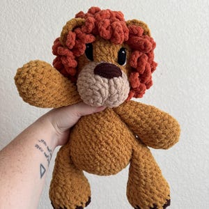 May include: A handmade crochet lion plush toy in shades of brown and orange. The lion has a fluffy, textured mane, a light brown snout, and dark brown accents on its paws. The toy is soft and cuddly, perfect for children.