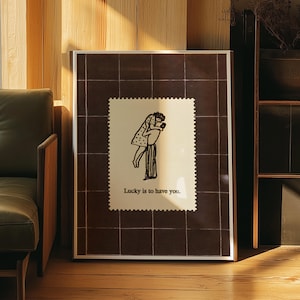 Puede incluir: Impresión artística enmarcada con una ilustración en blanco y negro de una pareja abrazándose. La obra de arte está sobre un fondo crema con el texto "Lucky is to have you." El marco tiene un acabado plateado y un patrón de cuadrícula marrón.