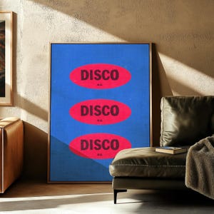 May include: A framed art print with the word "DISCO" in red, repeated three times on a blue background. The artwork is displayed in a room with a leather couch and a neutral-colored wall.
