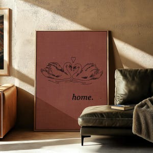 May include: Framed wall art featuring a pair of swans with intertwined necks and a heart above them. The artwork is on a reddish-brown background with the word "home" below the swans. The frame is a warm brown color.