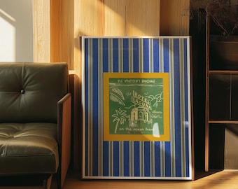May include: Framed vintage-style poster with blue and white striped background. The poster features a green and yellow central design with the words "HOTEL LAGUNA" and "on the ocean front". The poster is leaning against a wooden wall.