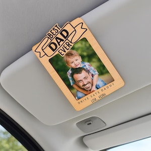 Könnte beinhalten: Ein hölzerner Sonnenblendenclip für Autos mit einem rechteckigen Rahmen, der ein Foto eines Mannes und eines Kindes enthält. Auf der Oberseite des Clips steht in schwarzer Schrift "BEST DAD EVER". Unten steht "DRIVE SAFE DADDY LOVE, HORN".
