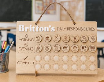 May include: Wooden daily responsibility chart with the name "Britton's" in white letters. The chart includes illustrated icons for morning and evening tasks, with a section for completed tasks. A rope handle is attached at the top.