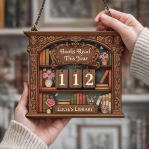 May include: A wooden sign shaped like a bookshelf, featuring the text "Books Read This Year" and the number "112". The sign also includes the text "Lucie's Library". The sign is brown with floral and book details.