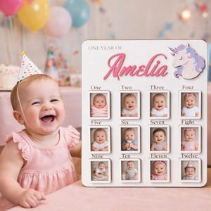 Peut inclure: Un tableau photo en bois blanc avec le nom "Amelia" en écriture rose, comportant douze petits cadres photo pour chaque mois de la première année d'un bébé. Un motif de licorne est en haut à droite. Un bébé en robe rose et chapeau de fête sourit.