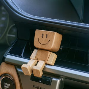 May include: A wooden figure with a smiling face carved on its head. The light brown figure sits on a car's dashboard vent. The figure's arms and legs are jointed, allowing for movement. The car's interior is visible in the background, with a start/stop button.