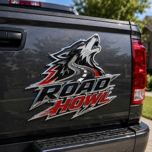 May include: A metallic emblem adhered to a dark gray truck tailgate featuring a stylized howling wolf graphic integrated with a road design and the text ROAD HOWL in bold black and red lettering with a silver chrome outline and finish.