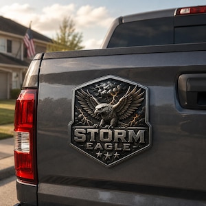Op de afbeelding: Een donkergrijs metalen embleem bevestigd op de achterklep van een pick-up truck. Het embleem is schildvormig en bevat een reliëf van een adelaar met gespreide vleugels, donkere onweerswolken, bliksemschichten, de tekst STORM EAGLE en drie sterren.