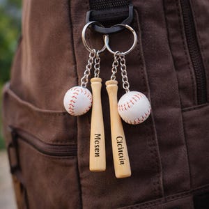 Könnte beinhalten: Zwei Baseball-Schlüsselanhänger hängen an einem schwarzen Verschluss. Jeder Schlüsselanhänger zeigt einen kleinen, weißen Baseball mit roten Nähten und einen Miniatur-Baseballschläger aus Holz. Die Schläger sind mit Namen graviert. Der Hintergrund ist ein brauner Rucksack.