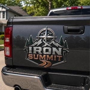 Op de afbeelding: De achterklep van een donkergrijze pick-up truck met een metalen embleem met een kompasroos, bergtoppen en dennenbomen. Het embleem toont de tekst IRON SUMMIT in dikke, verhoogde letters met een gebogen wegafbeelding onderaan.