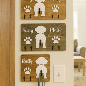 May include: Three wooden dog leash holders with a dog silhouette, paw prints, and hooks. The top two holders have the names "Rusty" and "Maisy". The bottom holder has the name "Rusty". The holders are mounted on a cream wall.