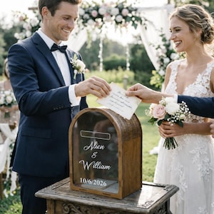May include: A wooden wedding card box with a clear front, inscribed with "Alien & William" and the date "10/6/2026". The box is placed on a table, and a person is inserting a card into the slot. A bride and groom are in the background.