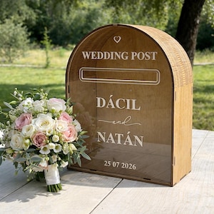 May include: Wooden wedding post box with the words "WEDDING POST" and the names "DÁCIL and NATÁN" and the date "25 07 2026" engraved on the front. A bouquet of white and pink roses and other flowers is next to the box.