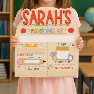 May include: Wooden sign with the name "SARAH'S" in red letters. The sign is decorated with colorful illustrations and text, including "FIRST DAY OF," "DATE," "I AM," "WHEN I GROW UP," "THINGS I LOVE," and "MY TEACHER IS."