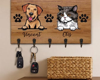Puede incluir: Un llavero de madera con un golden retriever y un gato gris y blanco. Los nombres 'Vincent' y 'Otis' están debajo. Huellas de patas negras y ganchos para llaves están debajo. Las llaves cuelgan de los ganchos. Una cesta tejida está debajo.