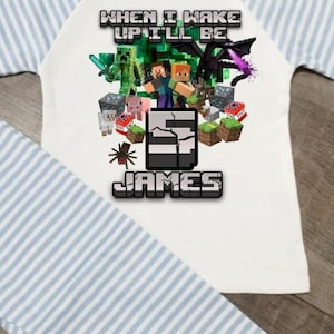 May include: A white t-shirt with light blue striped raglan sleeves featuring a Minecraft graphic print. The text reads WHEN I WAKE UP I'LL BE, followed by a large number 5 and the name JAMES, surrounded by various game characters and blocks.