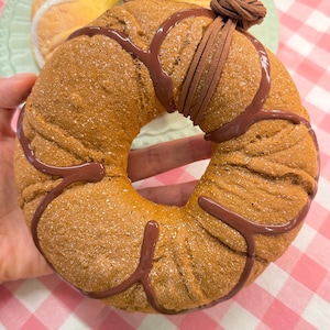 May include: A brown, ring-shaped pastry with a chocolate drizzle and a leather tie. The pastry is on a pink and white checkered surface. Another pastry with white icing is visible in the background.