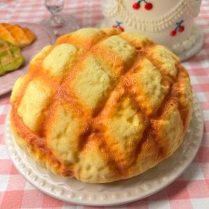 May include: A yellow and orange melon pan pastry sits on a white plate. The bread has a grid-like pattern and is surrounded by a reddish-orange color. Other pastries and a cake with cherries are in the background.