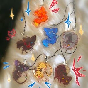 May include: A collection of seven clear acrylic charms featuring illustrated cartoon creatures in various colors including orange, blue, grey, purple, yellow, and brown, each attached to a black cord with a silver metal clasp and jump ring.