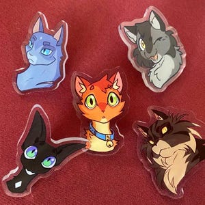 May include: Five clear acrylic cat-themed pins. The pins feature cartoon cat faces in various colors, including blue, orange, black, gray, and brown. Each pin has a unique expression and design, with details like collars and winking eyes.