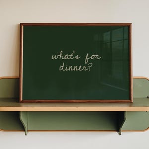 May include: A rectangular dark green framed sign with the white cursive text "what's for dinner?" resting on a sage green wall-mounted shelf with rounded edges and a light wood trim, featuring a clean design and a smooth, matte finish throughout.