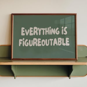 May include: A rectangular framed print with a dark green background and the white text "EVERYTHING IS FIGUREOUTABLE" in a rounded, bold font. The print is displayed on a horizontal, olive green wall-mounted shelf with light wood trim edges.