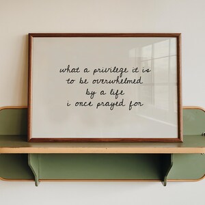 May include: A rectangular framed print with a dark wood border featuring the black text: what a privilege it is to be overwhelmed by a life i once prayed for. The print rests on a sage green wall-mounted shelf with rounded edges and light trim.