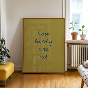 May include: A framed poster with the words "how lucky are we" in blue script on a yellow-green background. The poster is in a room with a yellow ottoman, a white couch, and plants.
