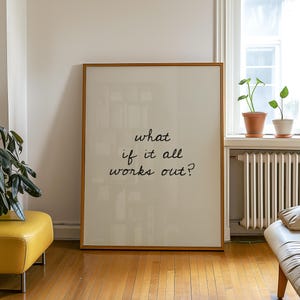 May include: A large framed print with the text "what if it all works out?" in black script. The frame is a light brown colour. The print is leaning against a white wall in a room with hardwood floors.