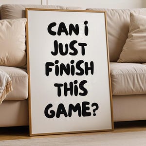May include: A framed art print with the text "CAN I JUST FINISH THIS GAME?" in black, bold letters. The print is displayed in a living room setting, leaning against a beige sofa. The room features neutral tones and wooden furniture.
