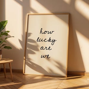 May include: A framed print with the words "how lucky are we" in black script. The artwork is displayed against a cream wall, with a small wooden side table and a potted plant in the foreground. The room is lit by natural light.