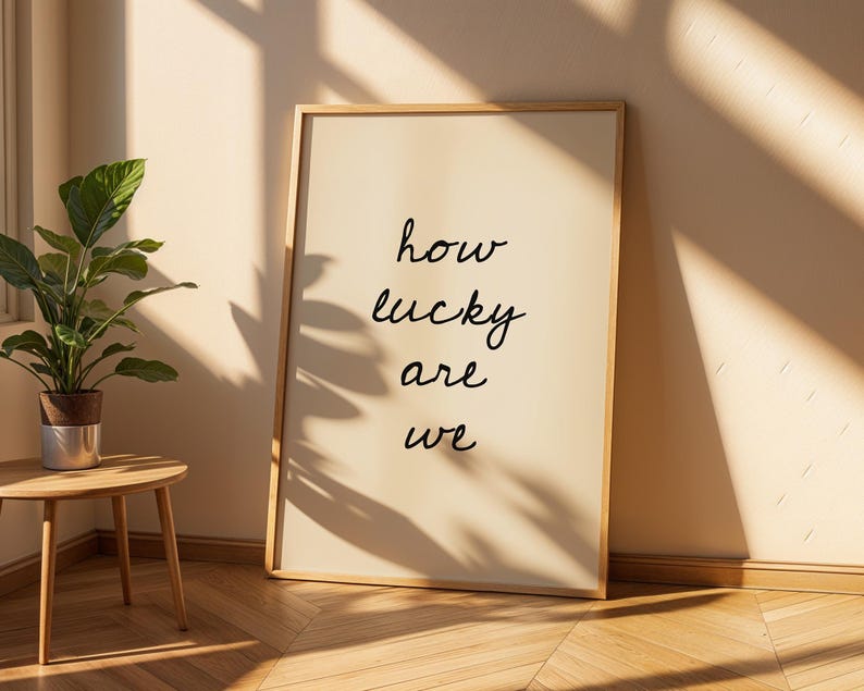 May include: A framed print with the words "how lucky are we" in black script. The artwork is displayed against a cream wall, with a small wooden side table and a potted plant in the foreground. The room is lit by natural light.