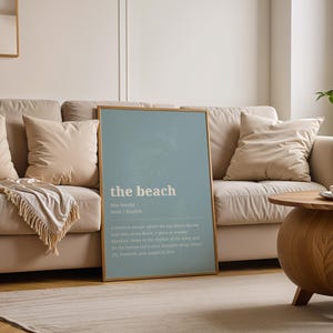Puede incluir: Un p&oacute;ster enmarcado con la palabra "the beach" en texto blanco sobre un fondo azul claro. El p&oacute;ster est&aacute; apoyado contra un sof&aacute; beige con cojines a juego. Tambi&eacute;n hay una mesa de centro de madera y una planta.