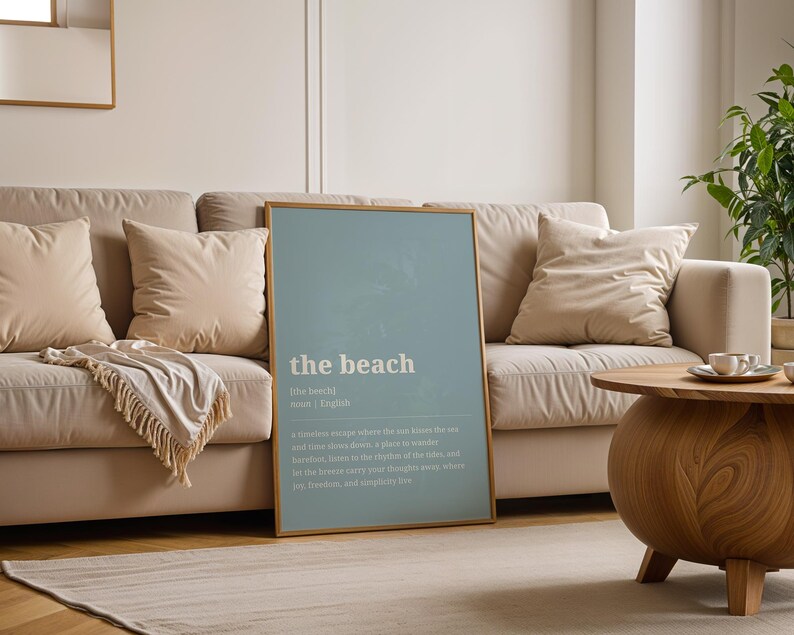 Puede incluir: Un p&oacute;ster enmarcado con la palabra "the beach" en texto blanco sobre un fondo azul claro. El p&oacute;ster est&aacute; apoyado contra un sof&aacute; beige con cojines a juego. Tambi&eacute;n hay una mesa de centro de madera y una planta.