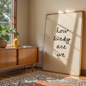 May include: A framed print with the words "how lucky are we" in black script leans against a wall. A wooden cabinet with a plant and a yellow vase sits near a window. A patterned rug covers the floor.