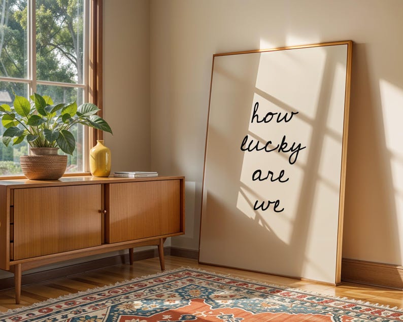 May include: A framed print with the words "how lucky are we" in black script leans against a wall. A wooden cabinet with a plant and a yellow vase sits near a window. A patterned rug covers the floor.
