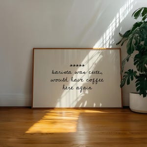 May include: A framed print with the text "barista was cute, would have coffee here again" in black script. The print has a cream-coloured background and a wooden frame. The print is displayed on a wooden floor.